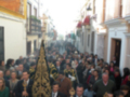 Una procesión religiosa en una calle de un pueblo con edificios blancos y azules. Las personas llevan trajes formales y mantas ornamentadas, con un hombre en el centro con una manto dorada.