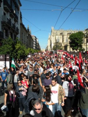 Una multitud en una manifestación, con banderas y carteles a la vista. En el fondo, edificios históricos y un edificio con torres se destacan. El cielo es claro y azul.