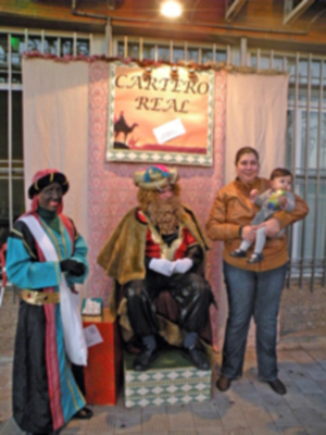 A tres personas vestidas con trajes tradicionales, posiblemente para una celebración navideña. En el fondo se puede ver un cartel que dice "CARTERERO REAL". La escena parece ser una representación o recreación de la tradición del reyes magos.