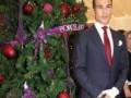 Un hombre en traje formal posa frente a un árbol de Navidad adornado con bolas rojas y purpuras. La decoración incluye una cinta que dice "Porcelanosa".