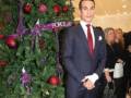 Un hombre en traje formal posa ante un árbol de navidad decorado con adornos rojos y morados. La imagen transmite un ambiente festivo navideño.
