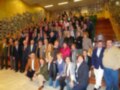 Una reunión formal de personas en un salón con paredes amarillas y escaleras. La mayoría están vestidos formalmente, posando para una foto en fila. En la pared hay muchas fotos y trofeos. La imagen transmite un ambiente de celebración o reconocimiento.