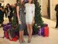 Dos mujeres posan frente a un árbol de Navidad decorado con regalos y cajas de presentación. La escena se desarrolla en un ambiente festivo, probablemente dentro de una tienda o centro comercial.