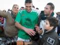 Un hombre en un jersey verde con una camiseta de fútbol, rodeado por personas que le están tomando fotos y hablando con él.