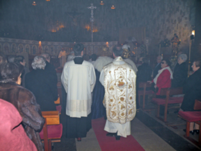 Una procesión religiosa en una iglesia, con personas vestidas de manera formal y un sacerdote en el centro. La imagen muestra una atmósfera serena y reverente, con luces suaves iluminando la escena.