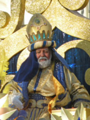 Un rey vestido con un traje dorado y azul, con un sombrero de copa y una corona en la cabeza. La imagen está iluminada con luces doradas y azules, creando un ambiente festivo.