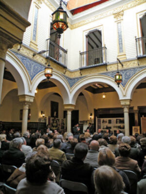Interior de un edificio histórico con arcos y columnas, gente sentada en filas escuchando a un hombre en el centro.