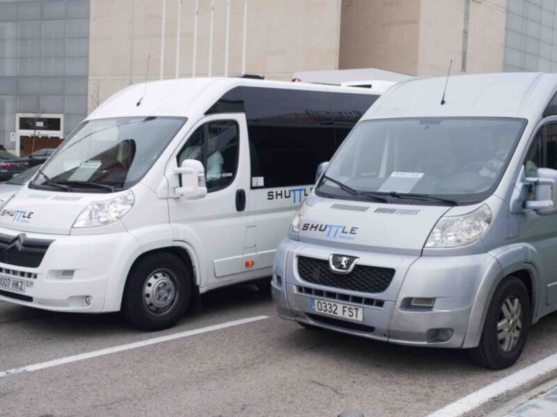 Dos vehículos blancos de la marca Shuttle estacionados en una calle con un edificio de fondo.