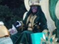 Person dressed in elaborate green and gold costume, seated on a throne-like chair, with a large, ornate hat with a red gem on top.