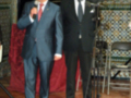 Dos hombres en trajes formales, uno hablando al micrófono y el otro escuchando. Fondo con mosaicos de azulejos.