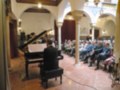 Un músico toca el piano en un salón con arcos y columnas, mientras otros espectadores observan la interpretación. La atmósfera es elegante y acogedora, con detalles arquitectónicos que resaltan la belleza del lugar.