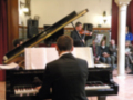 Un pianista en un concierto de música clásica, con una orquesta al fondo. El pianista toca un piano de cola y la orquesta incluye varios músicos, algunos tocando violines. La escena se desarrolla en un salón de conciertos con una columna y cortinas rojas.