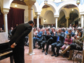 Un músico toca el piano en un salón con arcos y columnas, mientras una audiencia aplaude. La imagen muestra un ambiente formal con asistentes sentados en filas, algunos levantando las manos.