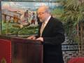 Un hombre con corbata y lentes habla en un podio frente a una pared de azulejos con imágenes históricas. A su lado, hay un escritorio con una hoja de papel y una silla roja. La escena parece ser un evento formal, posiblemente político o académico.