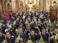 Imagen de una banda militar en uniforme, tocando instrumentos musicales tradicionales. El fondo muestra un altar con detalles dorados y una imagen religiosa. La banda está formada por hombres en trajes militares, con instrumentos como trompetas y tambores.