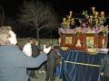 Un grupo de personas observa una procesión nocturna, con un carro de madera en el centro cubierto por una tela azul. La imagen muestra detalles como flores y figuras en el carro, con un hombre señalando hacia él. La escena se desarrolla al aire libre en una noche oscura, con un árbol sin hojas visible en el fondo.