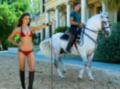 Una mujer en bikini rojo y botas altas posa junto a un hombre montando un caballo blanco en una playa.