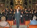 Una imagen de un coro en una iglesia, con un hombre en el centro dirigiendo a los cantantes. La decoración interna de la iglesia es elaborada, con arcos y vitrales. La escena parece ser una ceremonia religiosa o un concierto musical en una iglesia.