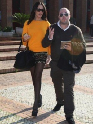 Una mujer con un vestido amarillo y pantyhose negros camina con un hombre en una calle adoquinada. La mujer lleva una bolsa de mano y el hombre sostiene un teléfono móvil. El fondo muestra una estructura de ladrillo y plantas verdes.