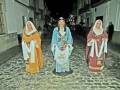 Tres personas vestidas con trajes tradicionales andaluces caminan por una calle adoquinada en la noche. La imagen muestra detalles de vestimentas típicas, con colores y diseños que reflejan la cultura local.