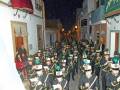 Desfile de bandas de música en una calle iluminada por la noche, con edificios tradicionales al fondo.