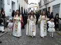 Un grupo de personas vestidas con trajes tradicionales blancos y dorados caminan por una calle adoquinada. Algunas portan sombreros negros, mientras que otras llevan velos blancos. La escena parece ser parte de una procesión o celebración cultural.