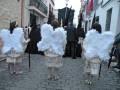 Tres figuras vestidas con trajes blancos y plumas, posiblemente representando ángeles, caminan por una calle adoquinada. Detrás de ellas, otros personajes con máscaras negras y trajes oscuros se desplazan. La escena parece ser parte de una procesión o celebración cultural, posiblemente relacionada con la Semana Santa.