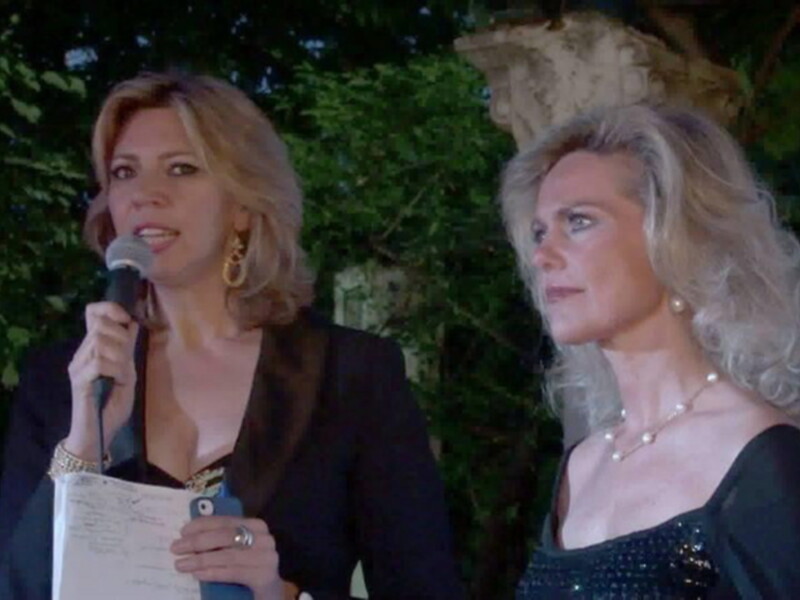 Dos mujeres en un evento al aire libre, una habla al micrófono mientras la otra escucha atentamente.