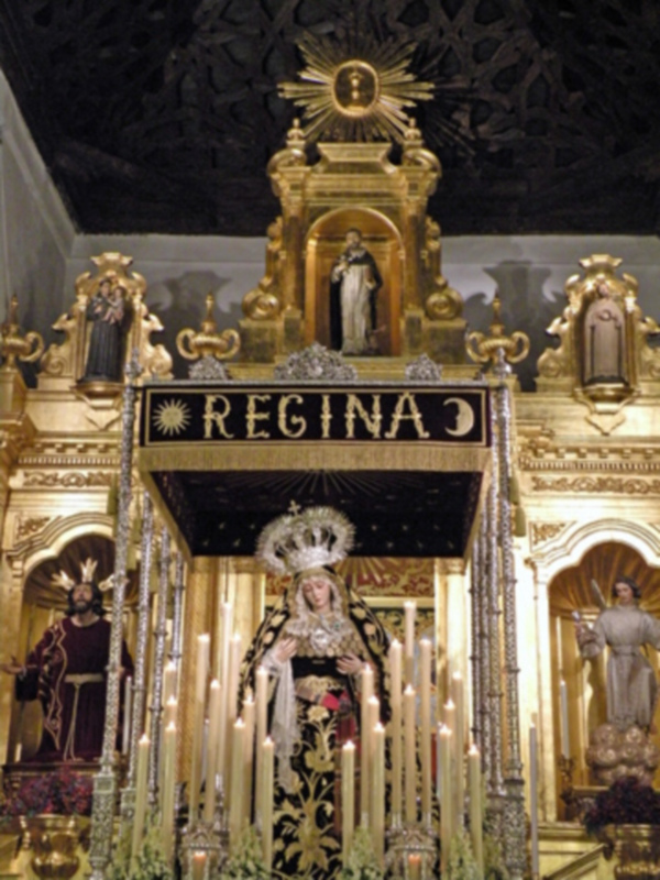 Un altar elaborado con estatuas de figuras religiosas, candelabros y adornos dorados. En el centro se encuentra una estatua de una figura vestida con un manto negro y dorado, rodeada de velas encendidas. La palabra "REGINA" se lee en letras doradas sobre el altar, sugiriendo que la figura central es la "Reina". El entorno es de estilo barroco, con detalles dorados y una atmósfera festiva.