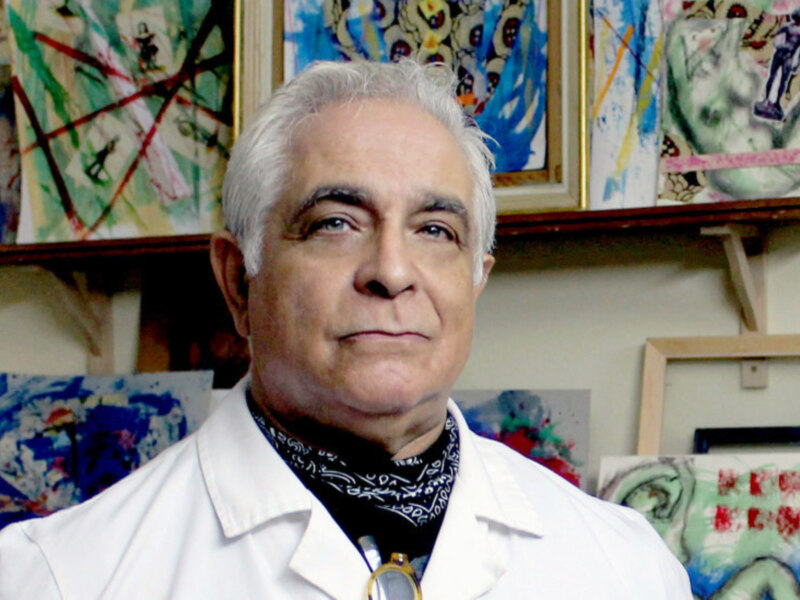 Un hombre con cabello blanco y barba, vistiendo una camisa blanca y un pañuelo negro con letras. El fondo es una pared llena de cuadros abstractos y coloridos.