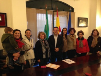 Una reunión formal en un salón con una mesa de madera, donde varias personas están posando para la foto. La imagen incluye a una persona con un niño en brazos, varias personas vestidas con abrigos y bufandas, y banderas de diferentes países en el fondo.