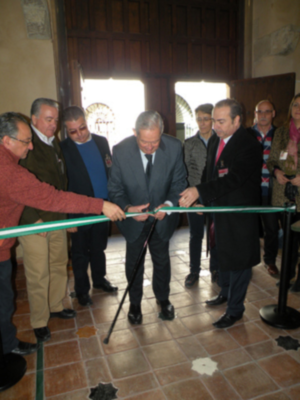 Un grupo de personas corta una cinta verde en un evento formal. El ambiente es interior, con paredes de madera y ventanas que permiten la entrada de luz natural. La cinta se encuentra en el centro, mientras los hombres usan trajes y chaquetas. La escena transmite una ceremonia o inauguración importante.