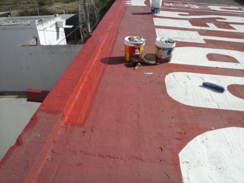 Un techo de color rojo con letras blancas pintadas. En el techo hay dos latas de pintura y un paño de limpieza, indicando que se está en proceso de pintar o restaurar el techo. La escena parece ser al aire libre, con un edificio blanco en el fondo y una valla de madera a la izquierda.