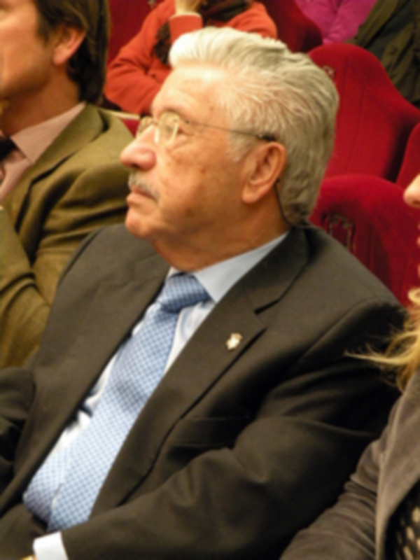Un hombre con barba y gafas, vestido de traje negro y corbata azul, sentado en un auditorio.