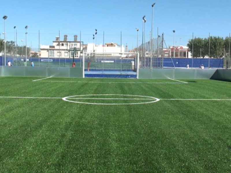 Campo de fútbol con césped artificial y portería, con edificios y árboles al fondo bajo un cielo azul claro.