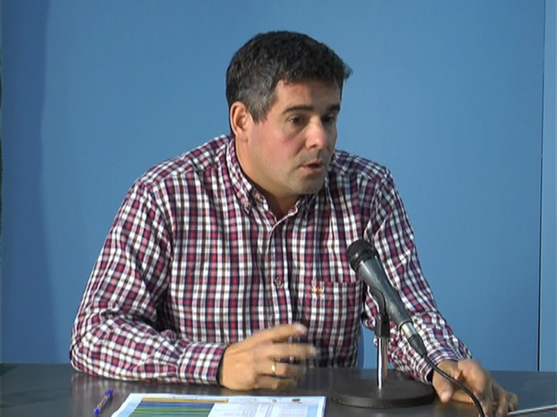 Persona hablando en una rueda de prensa con un micrófono y documentos en la mesa.