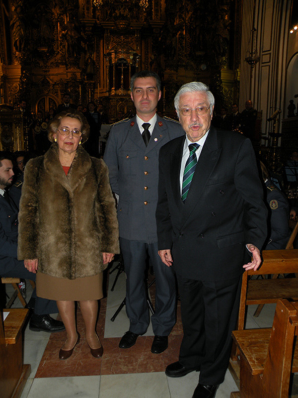Una familia posa en un ambiente decorado con una iglesia o templo en el fondo. La abuela está vestida con un abrigo de piel, mientras que los padres y el abuelo llevan trajes formales. La escena transmite un sentimiento de orgullo familiar y tradición.