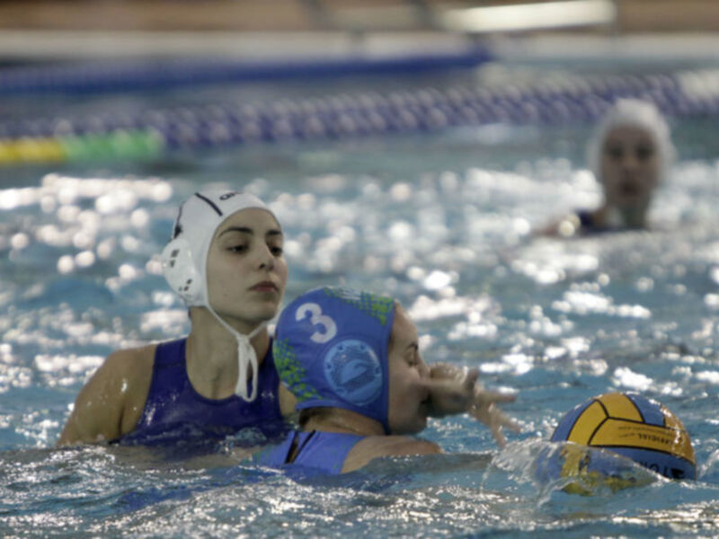 Una jugadora de waterpolo con un casco blanco y azul, vestida con una camiseta azul marino, lucha por la pelota en el agua. La imagen muestra a otra jugadora con un casco amarillo y azul en segundo plano. El agua es cristalina, reflejando la luz del día.