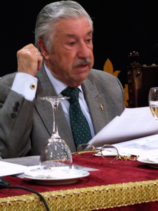 Un hombre en traje gris con corbata verde y una insignia en el pecho está sentado a la mesa de un evento formal. En frente de él hay una copa vacía y un plato con un cubilete de cristal. La mesa está cubierta con una manta roja y dorada, y hay un micrófono en el extremo izquierdo de la imagen.