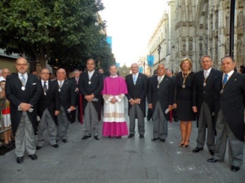 Grupal de personas en trajes formales y un sacerdote en el centro, posando frente a una catedral gótica.