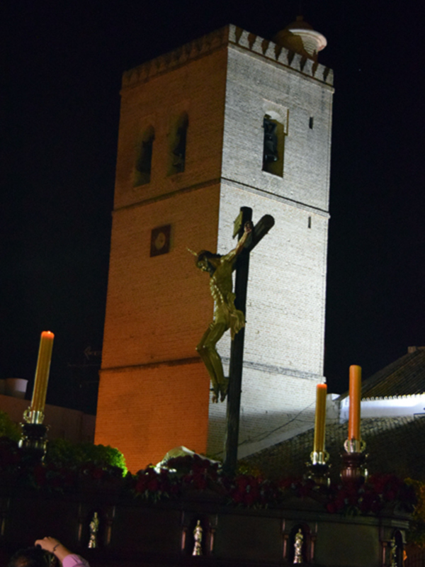 Una escena nocturna con un crucifijo iluminado en el centro. En el fondo, se puede ver una torre de ladrillo con detalles arquitectónicos. A la izquierda, hay una figura humana que parece estar tomando una foto. La escena está iluminada por faroles y la torre, que destaca en el cielo oscuro.