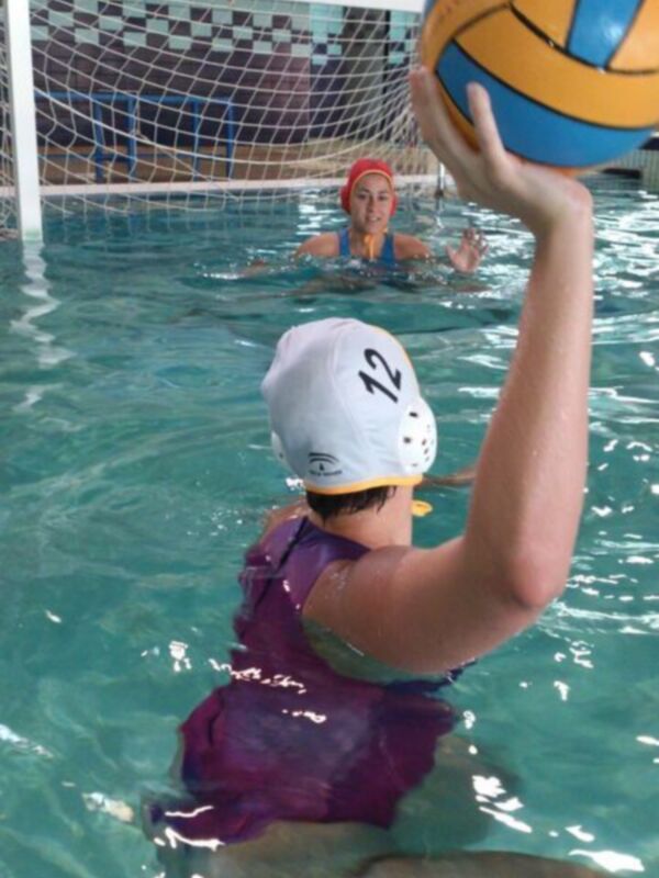 Un niño en un traje de baño azul y una gorra roja sostiene una pelota de voleibol de agua en un estadio de natación. La imagen muestra a otro niño en el fondo, también con una gorra roja y un traje de baño azul. La piscina tiene agua transparente con pequeñas burbujas y el techo de la sala está decorado con azulejos en colores pastel.