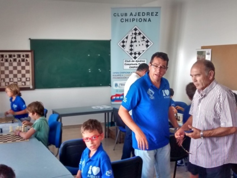 Un grupo de niños y adultos juegan al ajedrez en una sala con carteles informativos sobre el Club Ajedrez de Chipiona.