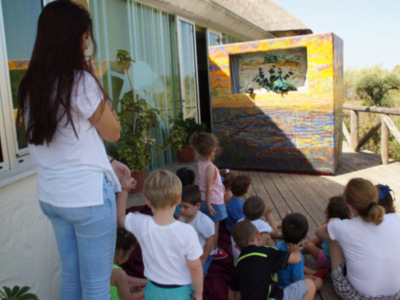 Una escena en un balcón de una casa con una mujer enseñando a niños pequeños. Los niños están sentados en el suelo, mirando hacia una pantalla de televisión colgada en la pared. La pantalla muestra un paisaje pintado con colores vibrantes, probablemente una escena de naturaleza. La casa tiene ventanas y un techo de paja, y el entorno parece ser una zona rural o semi-rural.