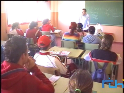 Clase de matemáticas en un aula con estudiantes sentados en sus mesas, el profesor está al frente enseñando. La pizarra contiene ecuaciones matemáticas y el logo de RTVE se puede ver en la esquina inferior derecha.