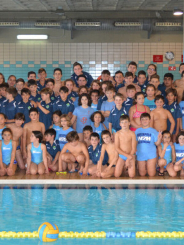 Grup de natació infantil a una piscina. Estan posant per a una foto grup al costat d'una piscina. Els nens i nenes estan vestits amb uniformes de natació. Al fons, hi ha una parella d'escorxadors i un calendari amb la data.