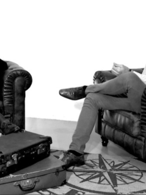 Dos hombres sentados en sillones de cuero, uno con una maleta junto a él. El suelo tiene un patrón circular y la pared es blanca.