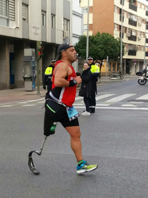 Un hombre con una prótesis corre por la calle, vestido con un chándal rojo y negro. Detrás de él, hay edificios urbanos con varios balcones y ventanas. La imagen muestra una calle con señales de tráfico, un ciclista en el fondo y vehículos a la distancia.