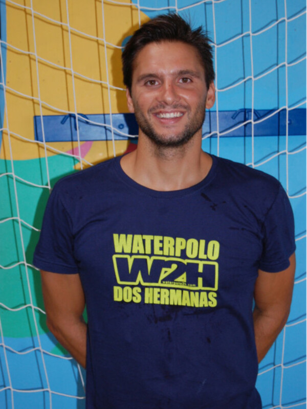 Un hombre sonríe frente a una pared de azulejos con un diseño de pelota de waterpolo. Lleva una camiseta azul con letras amarillas que dicen "Waterpolo W&H Dos Hermanas".