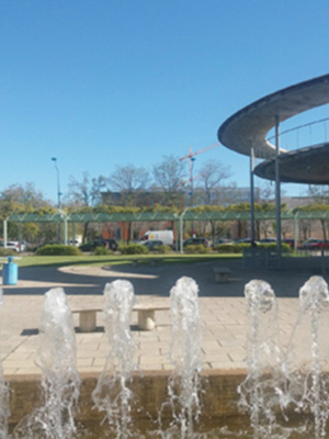 Fuentes de agua en un parque público con bancos y árboles al fondo. El cielo es azul claro sin nubes.